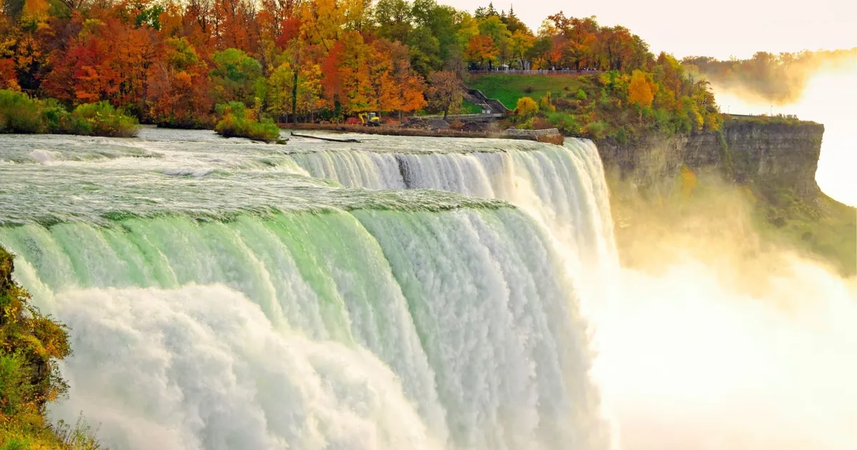 The Niagara River pouring over the brink of the falls with autumn trees on the bank above