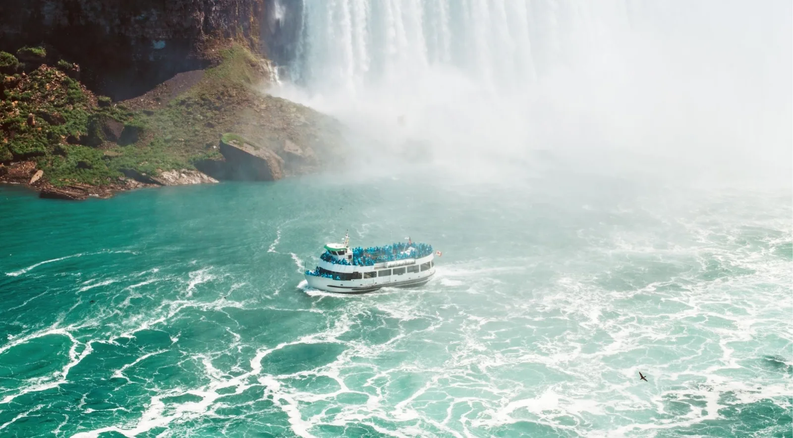 Tour boat crowded with passengers in blue ponchos turning in the emerald water at the foot of Horseshoe Falls