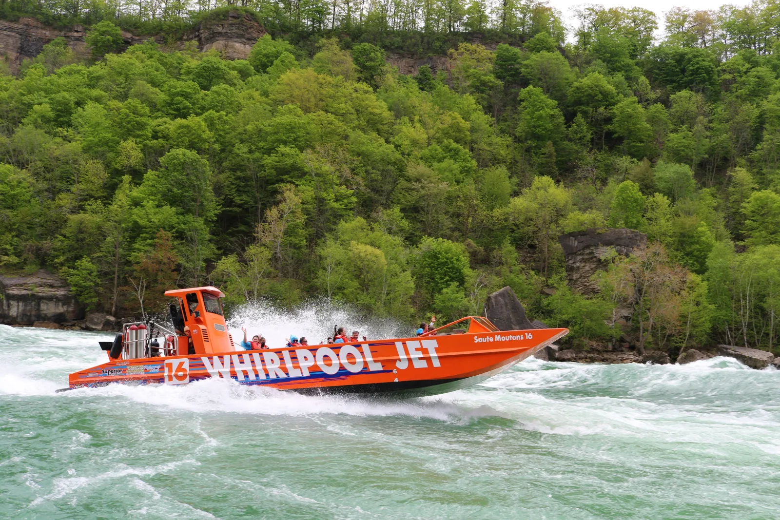 Orange Whirlpool Jet open-top boat running at speed through the rapids below the forested wall of the Niagara Gorge
