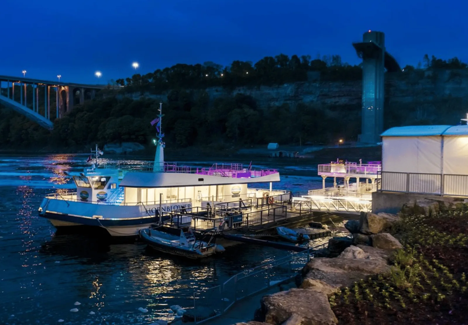 The Niagara City Cruises catamaran moored at its lit boarding dock at blue hour, the Rainbow Bridge and observation tower behind