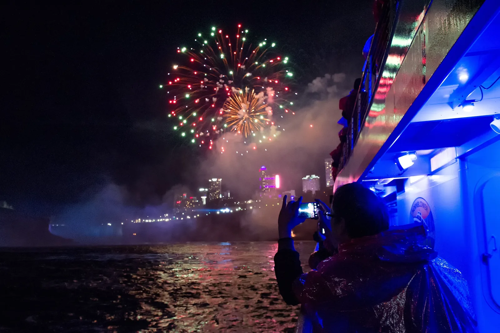 A passenger in a poncho filming fireworks from the boat rail, the illuminated Niagara Falls skyline glowing through the mist