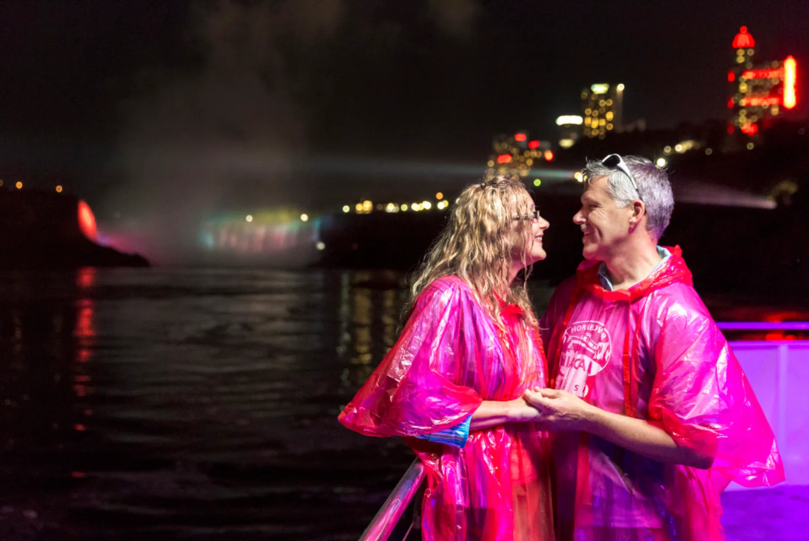 A couple in pink souvenir ponchos on deck at night, the colour-lit falls and city lights behind them