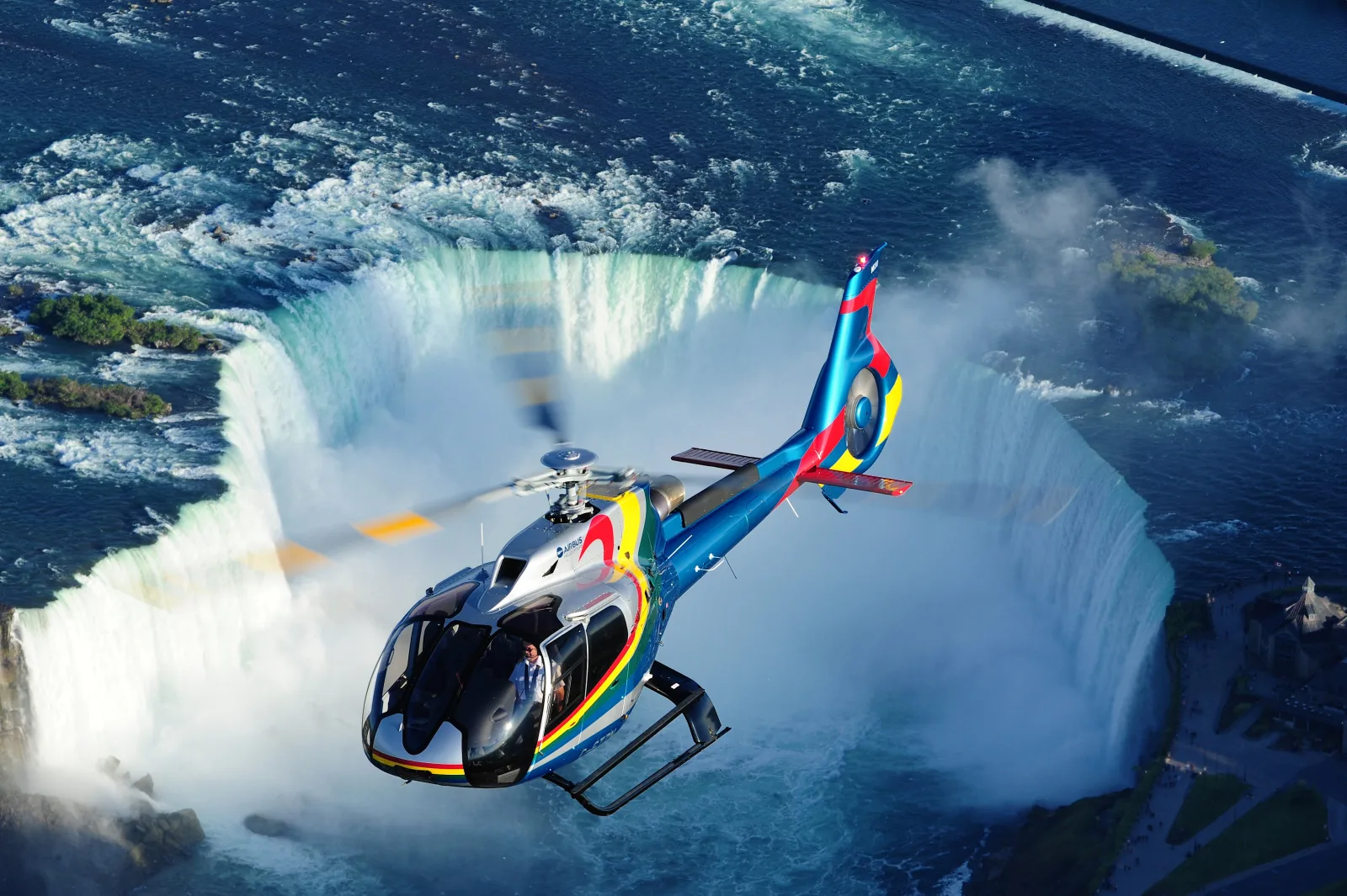 Brightly liveried Airbus H130 helicopter banking directly over the Horseshoe Falls, the full curve of the cataract and its mist below