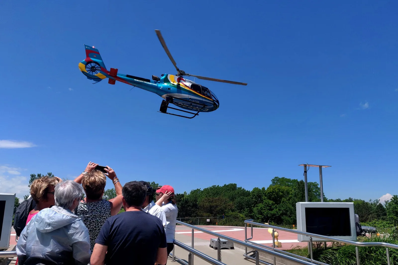 Waiting passengers photographing the Niagara Helicopters aircraft as it lifts off the Victoria Avenue heliport against a clear blue sky