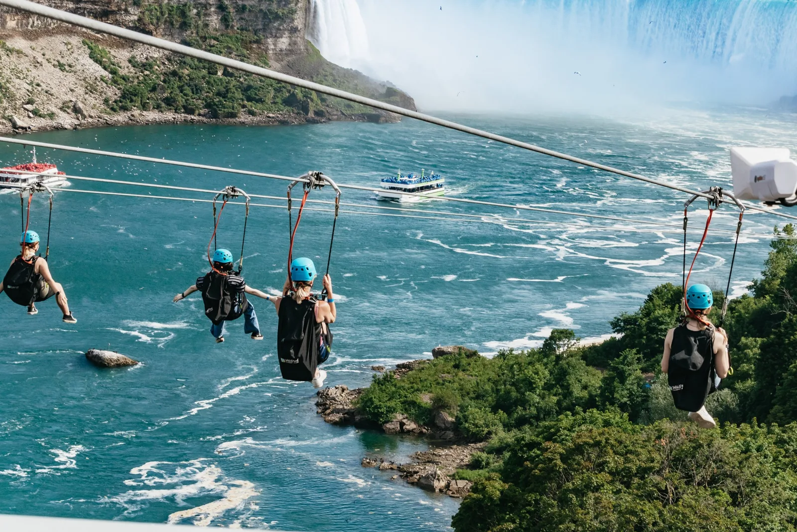 Four riders in blue helmets and black seated harnesses gliding down parallel ziplines above the Niagara River, Horseshoe Falls in mist ahead of them