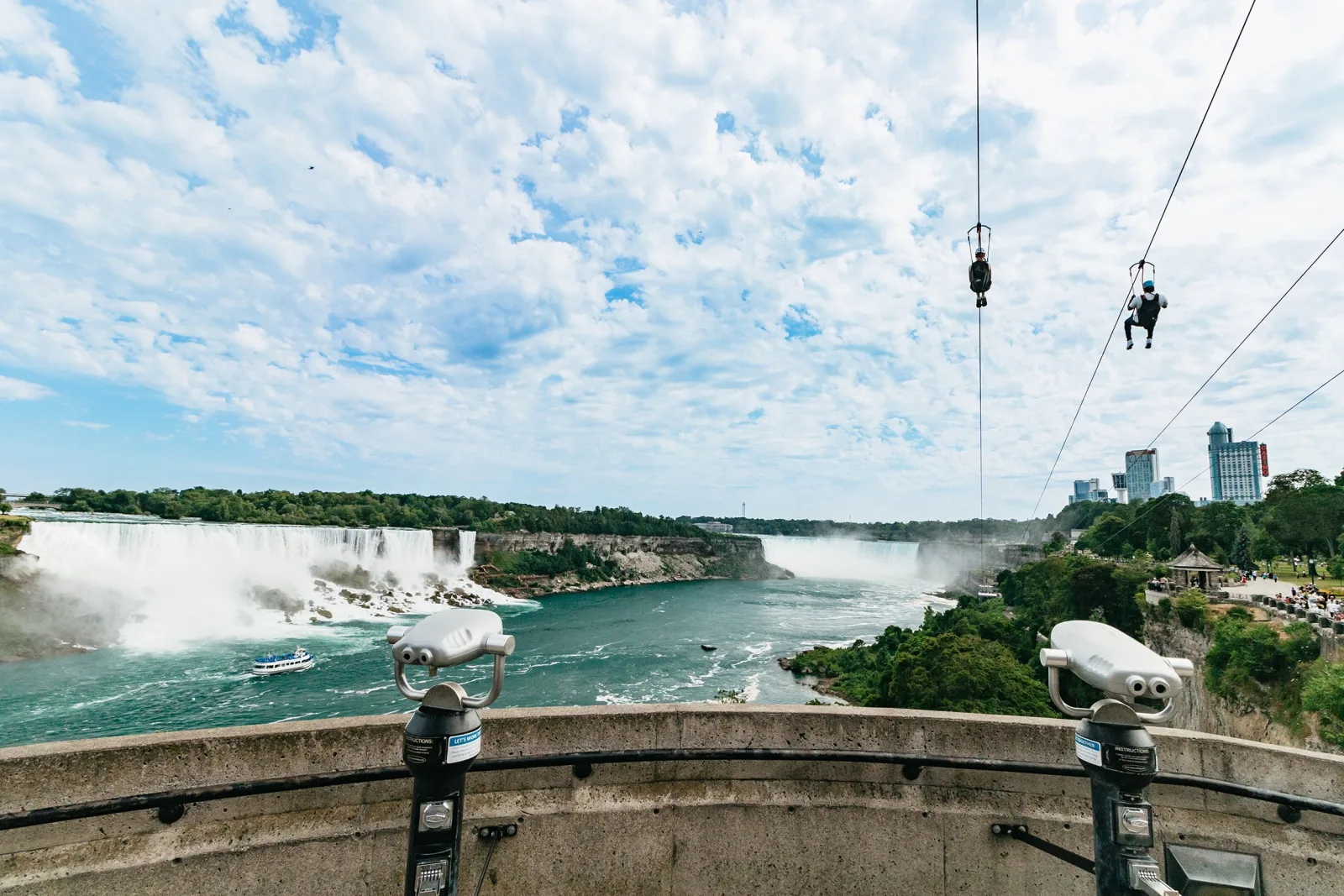 Two zipliners silhouetted against a cloud-filled sky above the gorge, the American Falls and a tour boat far below