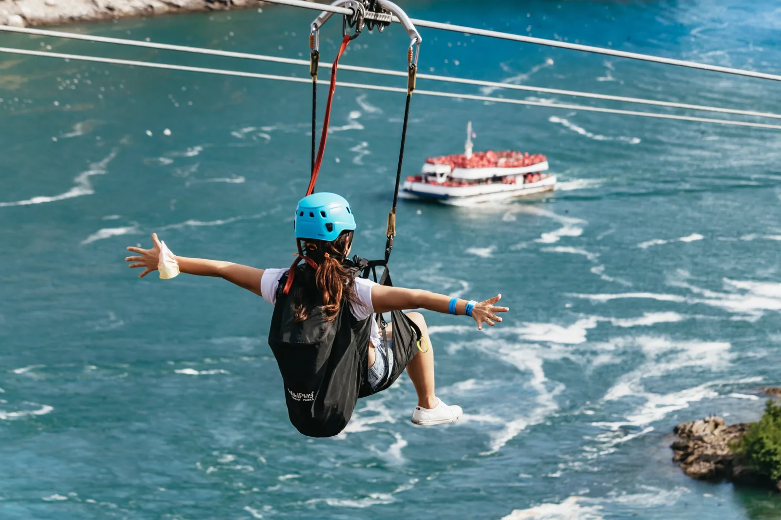 A rider with both arms outstretched, seated in the zipline harness high above the green Niagara River with a cruise boat passing beneath