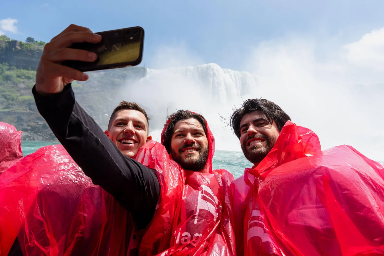Three friends in red ponchos photographing themselves on the boat deck with the American Falls and its mist behind them
