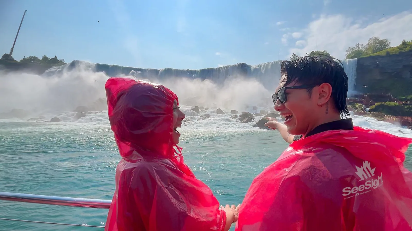 Two passengers in red ponchos laughing at the boat rail as the vessel passes the American Falls and its boulder field