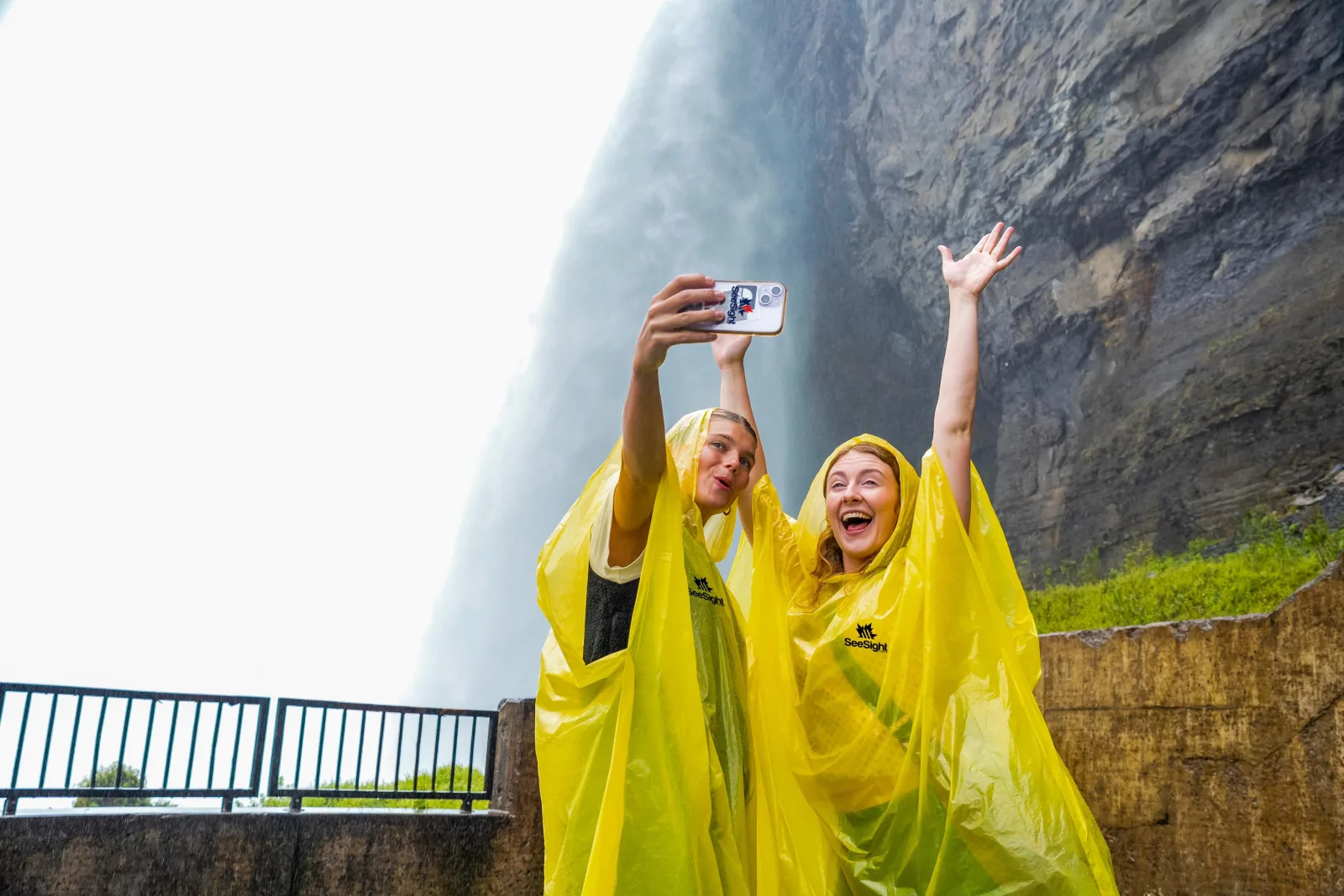 Two visitors in yellow ponchos on the lower observation deck, arms raised, the Horseshoe Falls curtain of water filling the frame behind