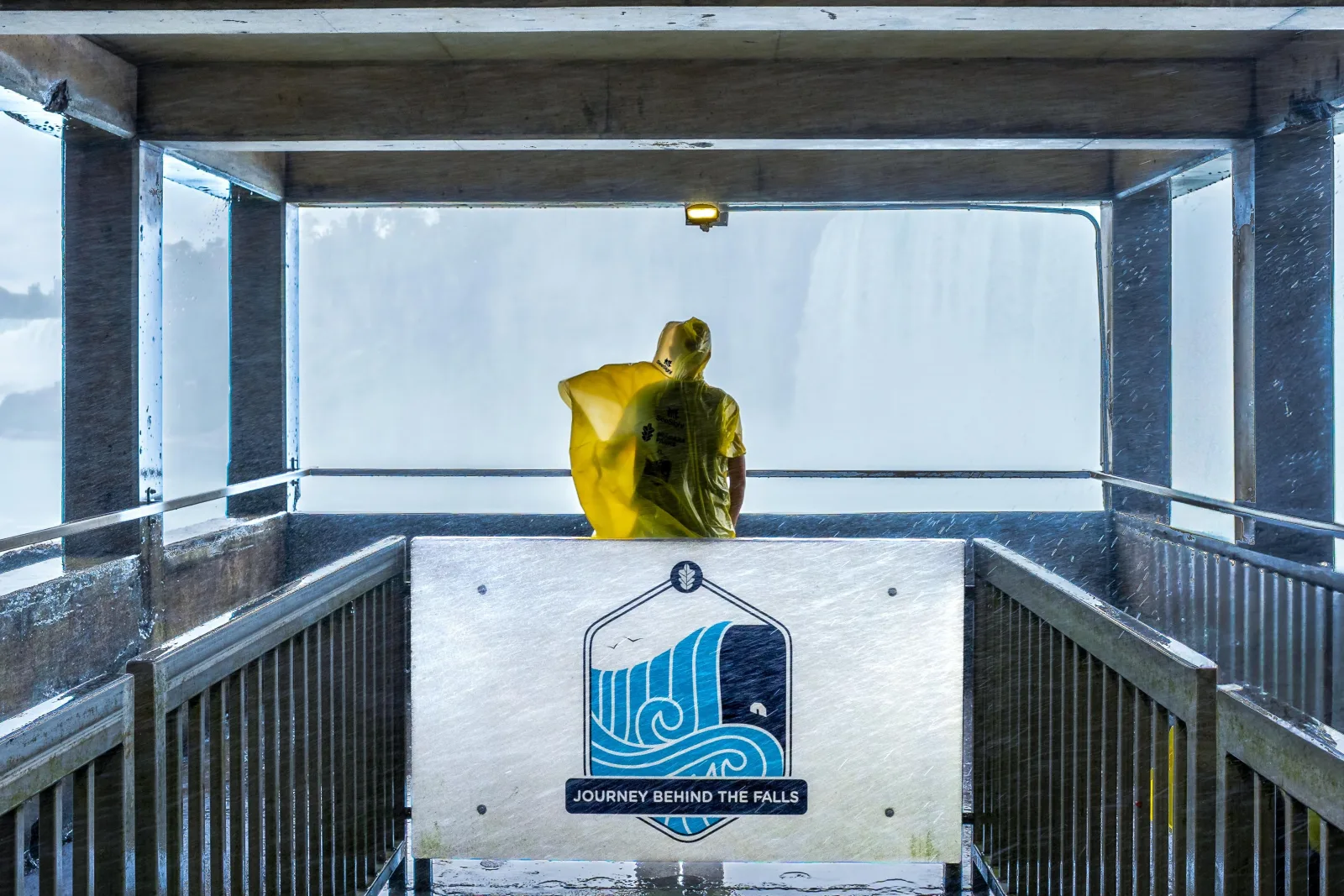A visitor in a yellow poncho at the Journey Behind the Falls lower deck, facing the sheet of falling water past a sign reading Journey Behind the Falls