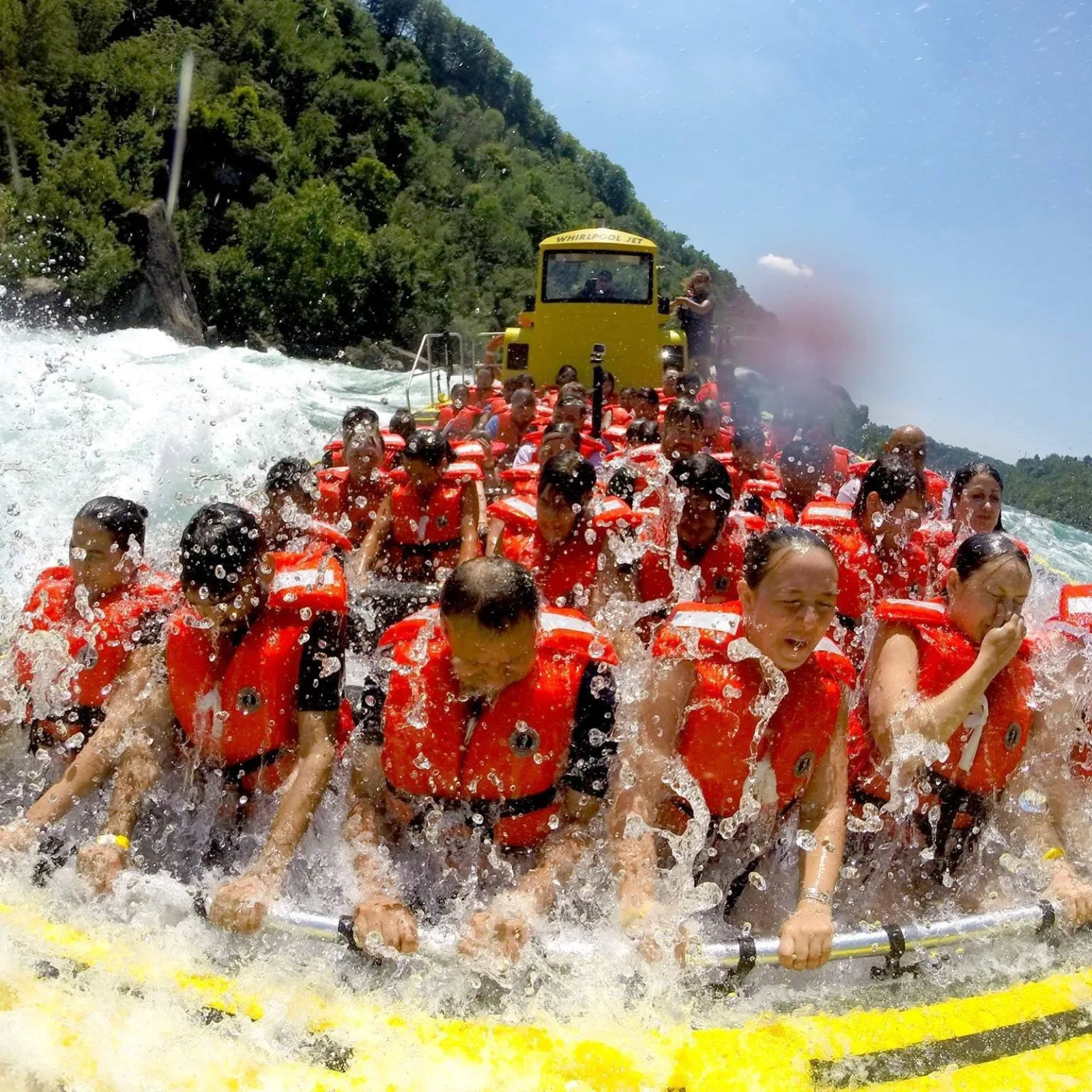 Riders in orange life jackets braced against the rail of a yellow Whirlpool Jet boat as a wall of Niagara River water breaks over the bow