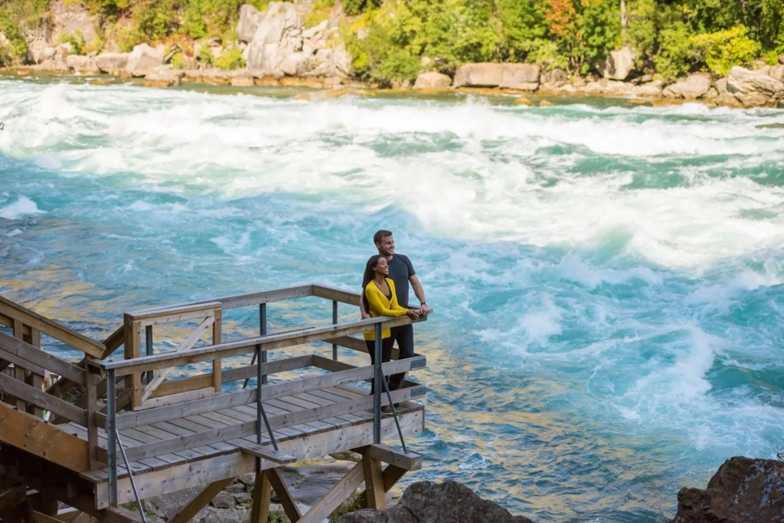 Two visitors on a timber viewing platform at river level, the Whirlpool Rapids breaking in turquoise and white just beyond the rail
