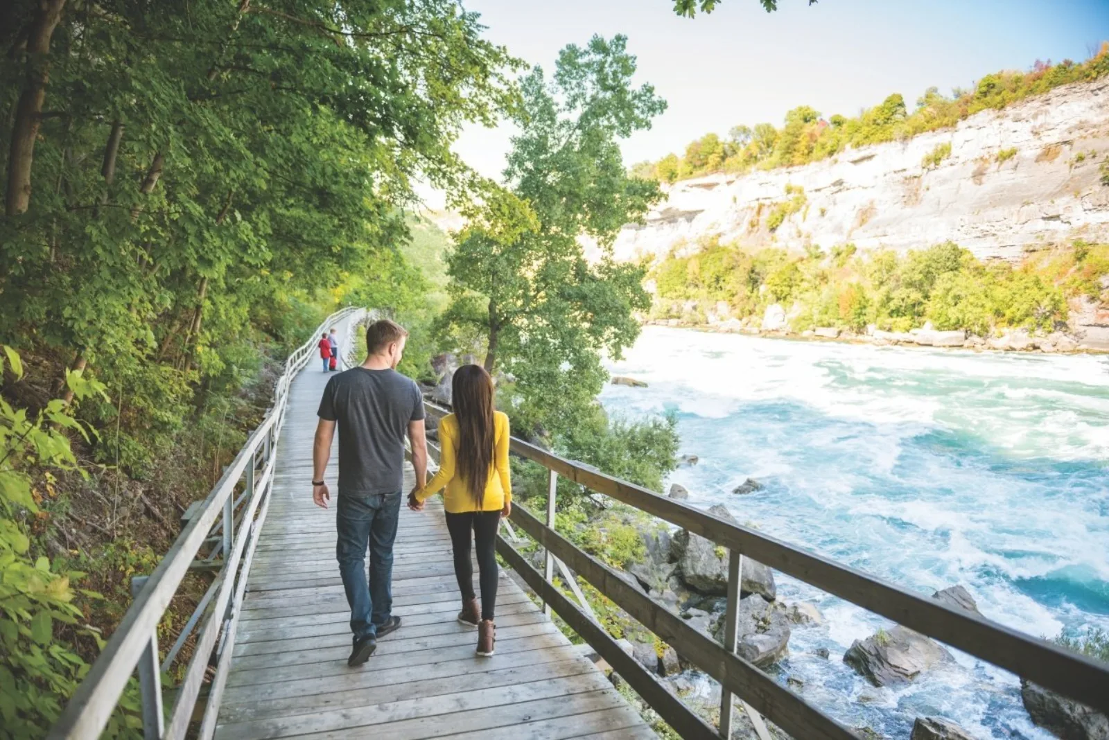 Visitors walking the level timber boardwalk deep in the Niagara Gorge, the pale limestone cliff face rising across the rapids